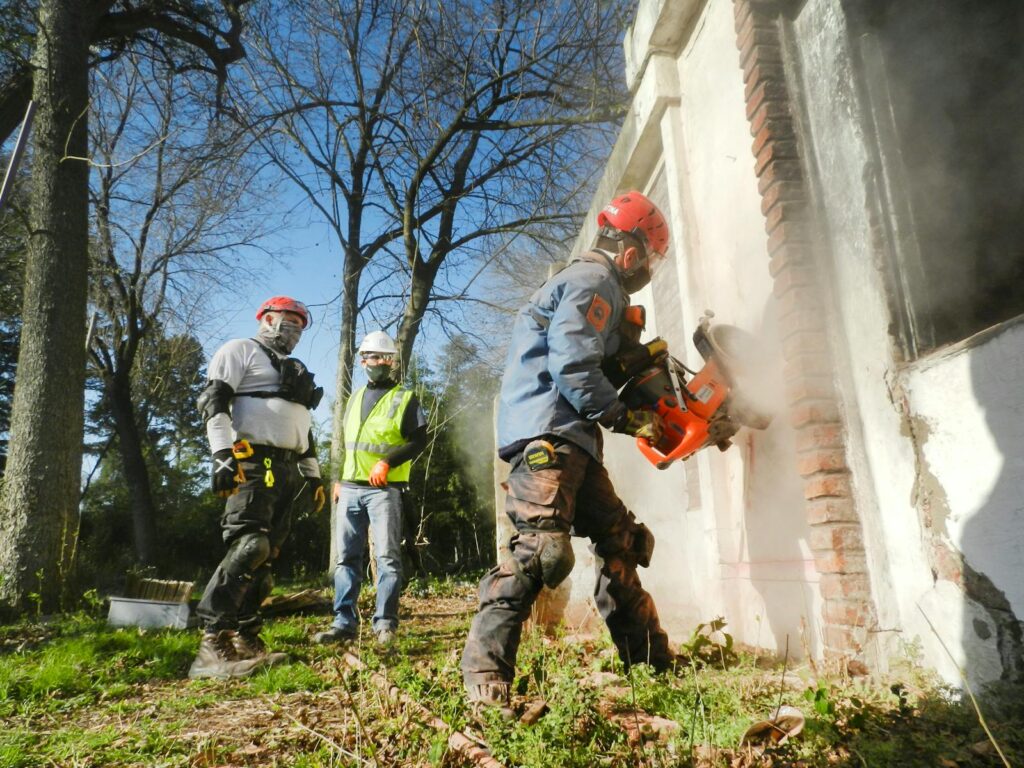Construction workers dressed in safety gear demolishing a brick wall with heavy machinery outdoors.
