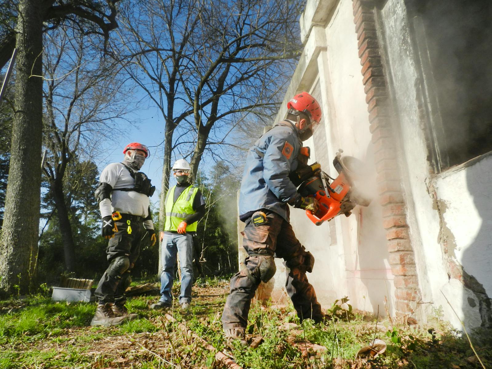 Construction workers dressed in safety gear demolishing a brick wall with heavy machinery outdoors.